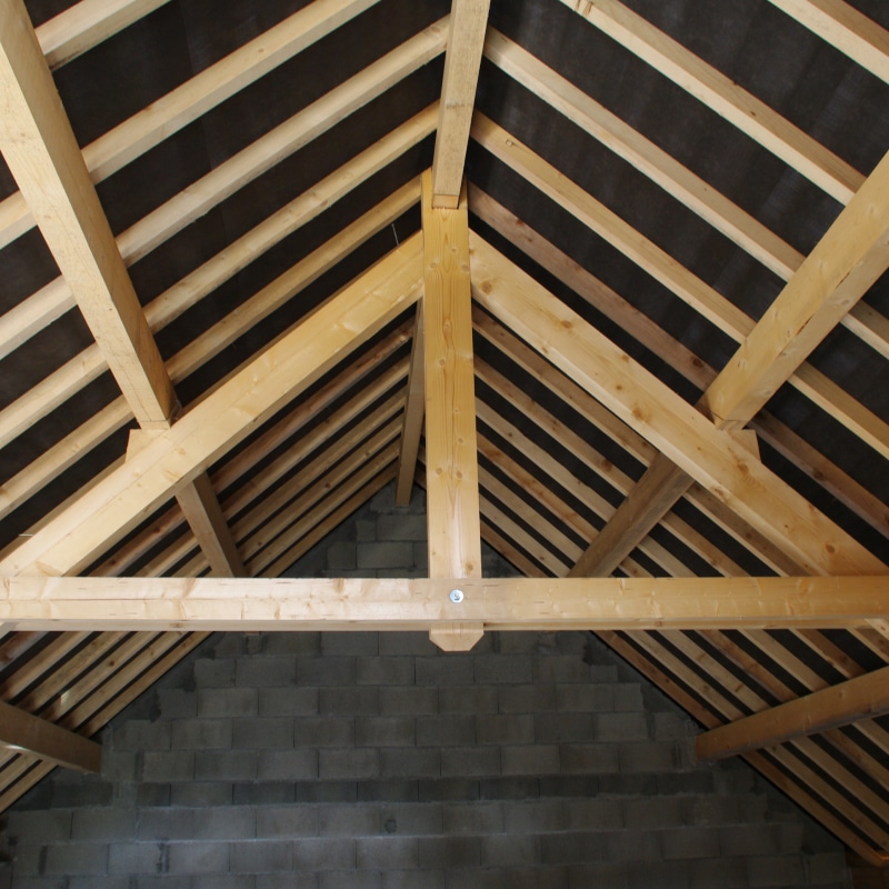 Charpente en bois d'une toiture en construction, avec chevrons et pannes apparents sur fond noir d'isolation et murs en blocs de béton gris.