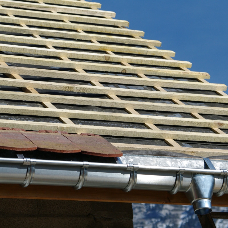 Toiture en construction, lattes de bois sur membrane noire, prête pour les tuiles. Nouvelle gouttière métallique installée sous des tuiles anciennes, ciel bleu.