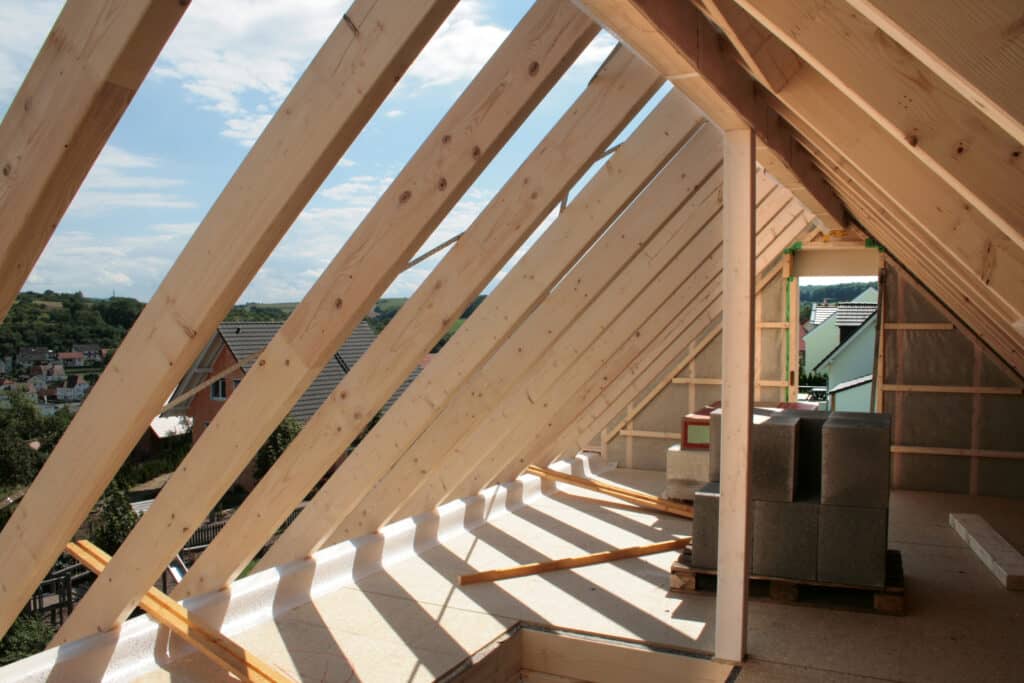 Grenier en Construction : Charpente Bois, Vue Village Intérieur de grenier en construction, charpente en bois apparente. Le sous-plancher est éclairé par le soleil, avec vue sur un village.