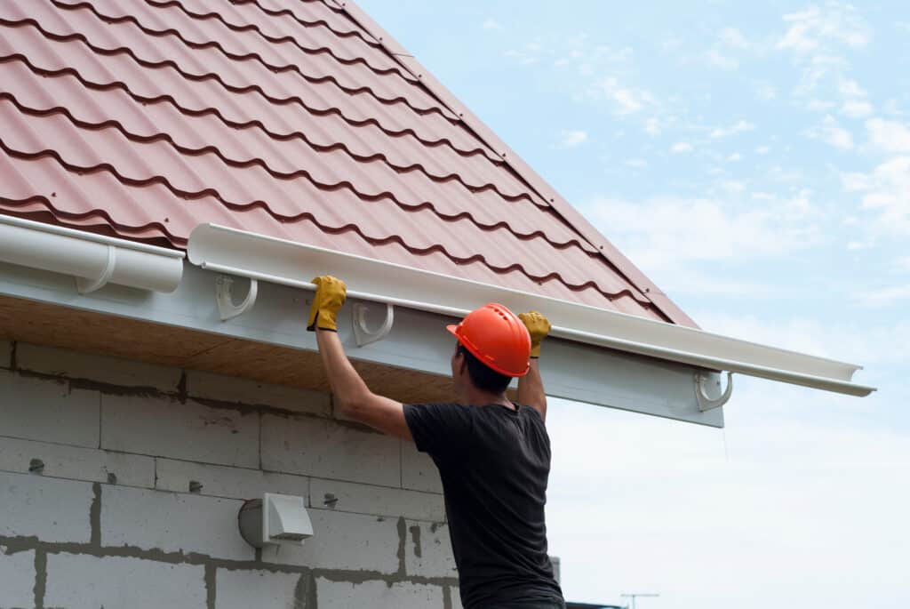 Couvreur en casque orange et gants jaunes installe une gouttière blanche sur le toit en tuiles métalliques rouges d'une maison.